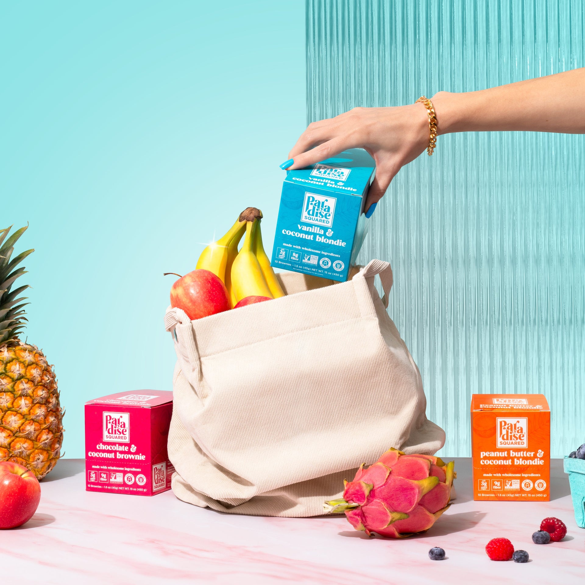 Hand lifting out box of Paradise Squared Vanilla & Coconut Blondies out of tan shopping bag. Surrounded by fruits like dragon fruit, apple, pineapple, raspberries, and blueberries. Light blue background.