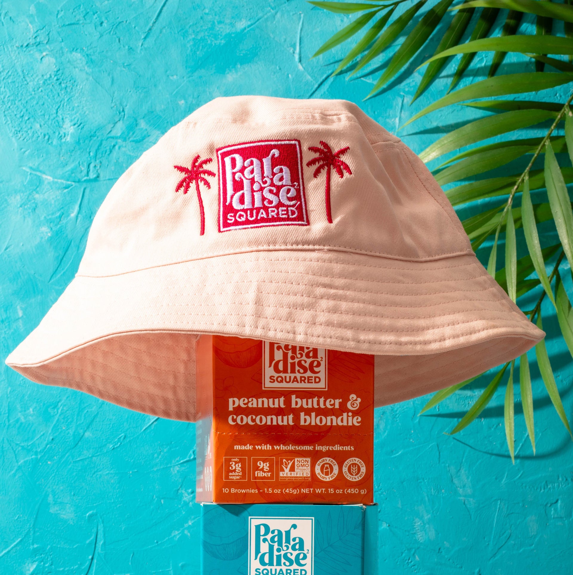 Light pink Paradise Squared bucket hat stacked on top of boxes of Paradise Squared. Bright blue background with palm fronds